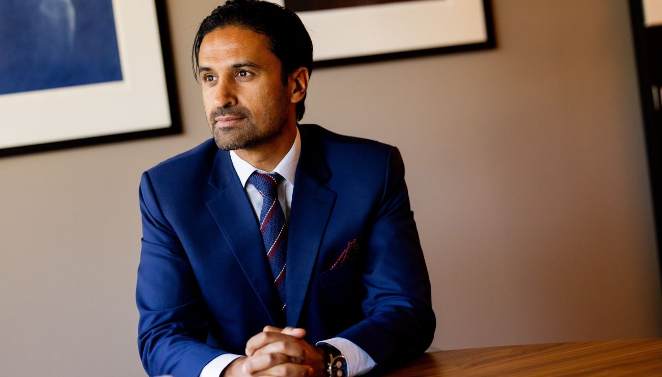 Formelt kledd mann som sitter i et kontor og fletter fingrene sammen på bordet foran seg.