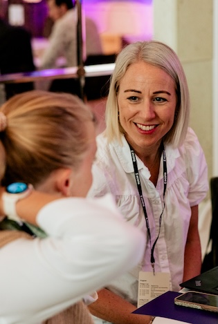 En kvinne med skulderlangt, lyst hår og hvit bluse smiler og snakker med en annen kvinne som er fotografert fra siden/bakfra, ved et registreringsbord med papirer og en mobiltelefon. Bakgrunnen er et rom med lilla belysning.