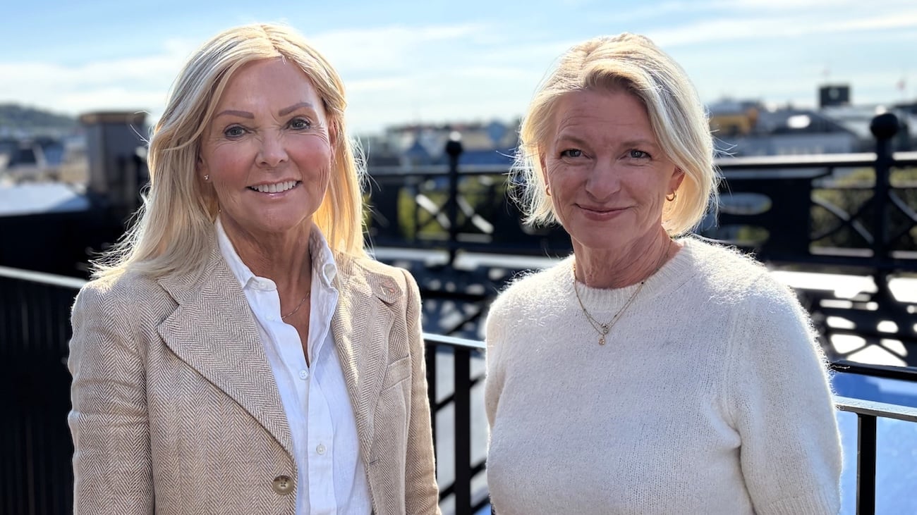 To kvinner smiler til kameraet på en takterrasse en solfylt dag, med byen i bakgrunnen.