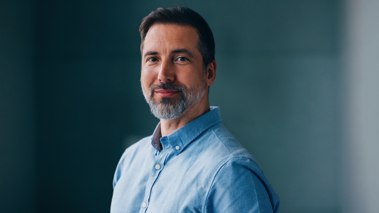 Kontorkledd mann ser i kamera og smiler mot blålig bakgrunn.