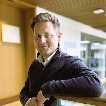 Mann som lener seg mot et glassgjerde mens han smiler lurt til kamera.