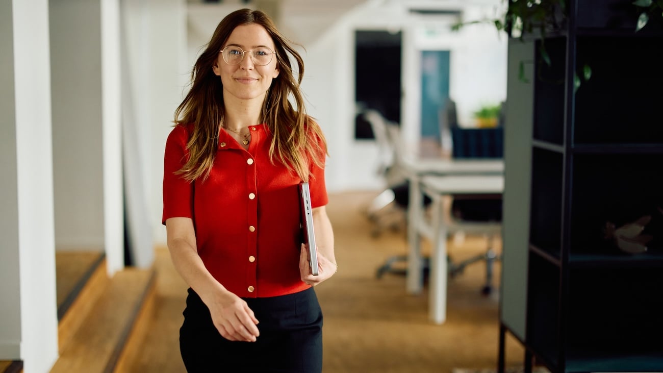 Ung kvinne i et kontorlandskap som kommer gående mot kamera med en PC under armen.