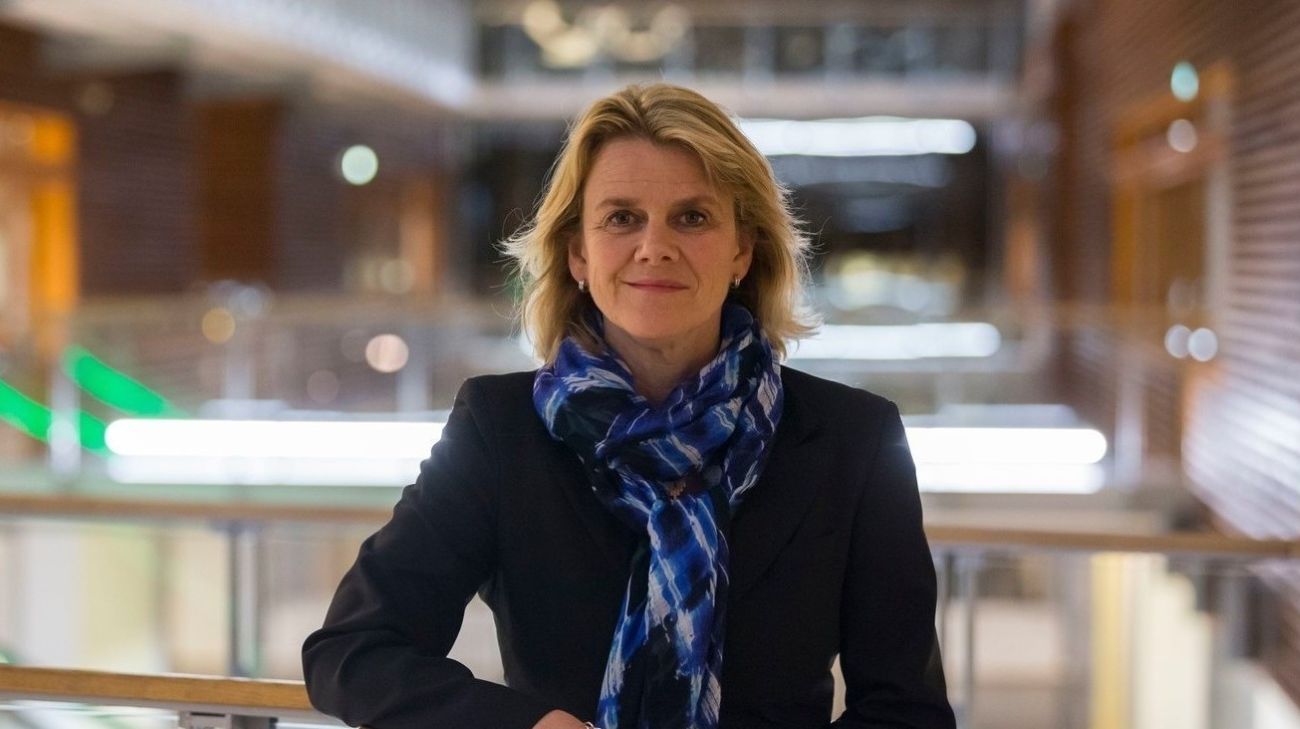 Kvinne med silkeskjerf rundt halsen som ser lurt bort på en annen person i en sal med mennesker.