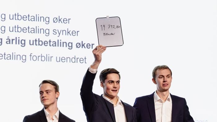 Tre unge menn som står på en scene og holder opp en tavle der de har skrevet svaret på utslagsspørsmålet under NM i økonomi 2026.