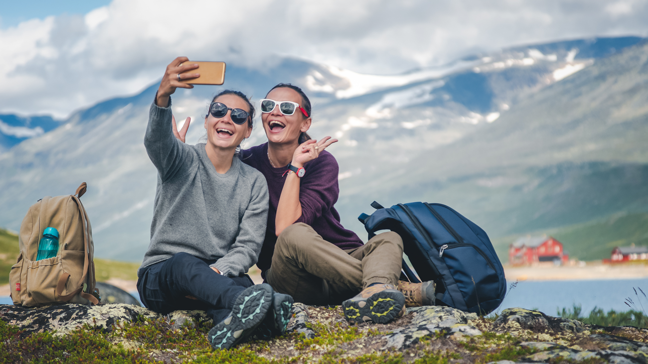 To kvinner som som smiler bredt mens de tar en selfie ute i naturen med fjor og fjell i bakgrunnen.