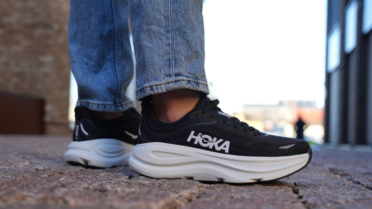 Beina til en person som står på brostein med et par nye, sorte HOKA joggesko.