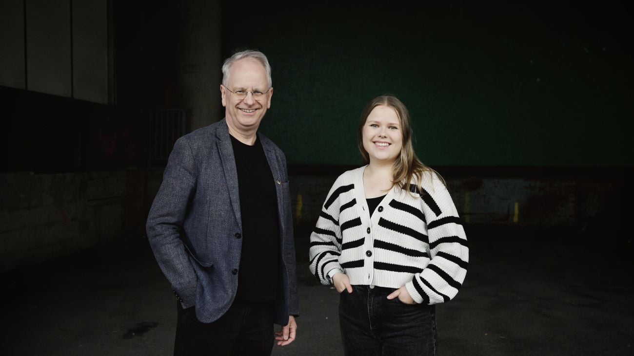 Far og datter som begge står med hendene i lommene og smiler i kamera.