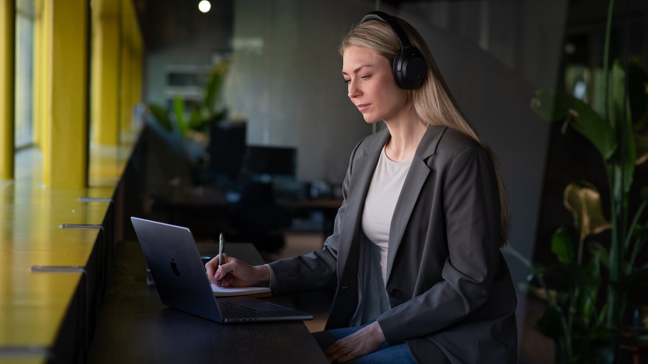 Profesjonell kvinne med hodetelefoner jobber fokusert foran laptop i moderne kontormiljø, tar notater for faglig utvikling.