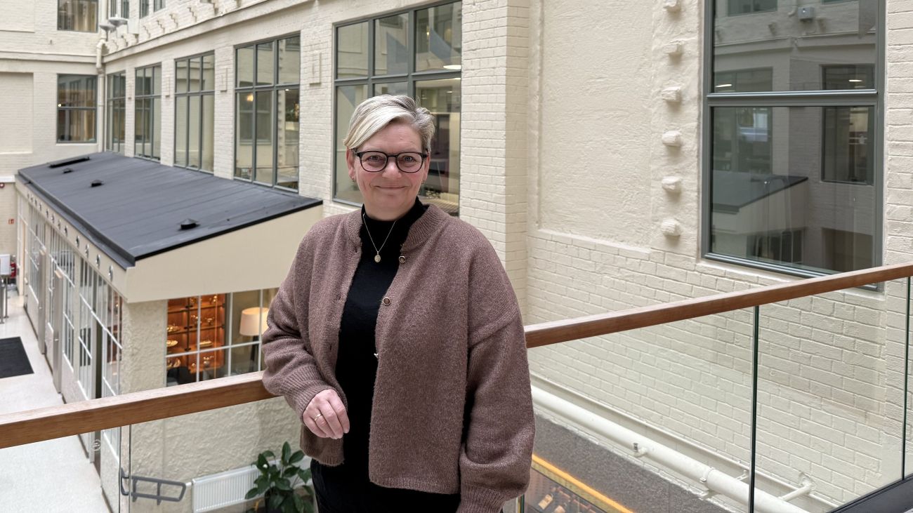 Kvinne som står og lener seg mot et glassrekkverk i en stor og lys bygning mens hun smiler forsiktig til kamera.