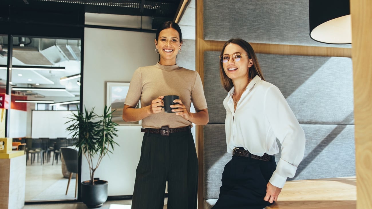To unge kvinner kledd i business casual antrekk som står sammen i et kontorlandskap i en hyggelig kaffeprat.