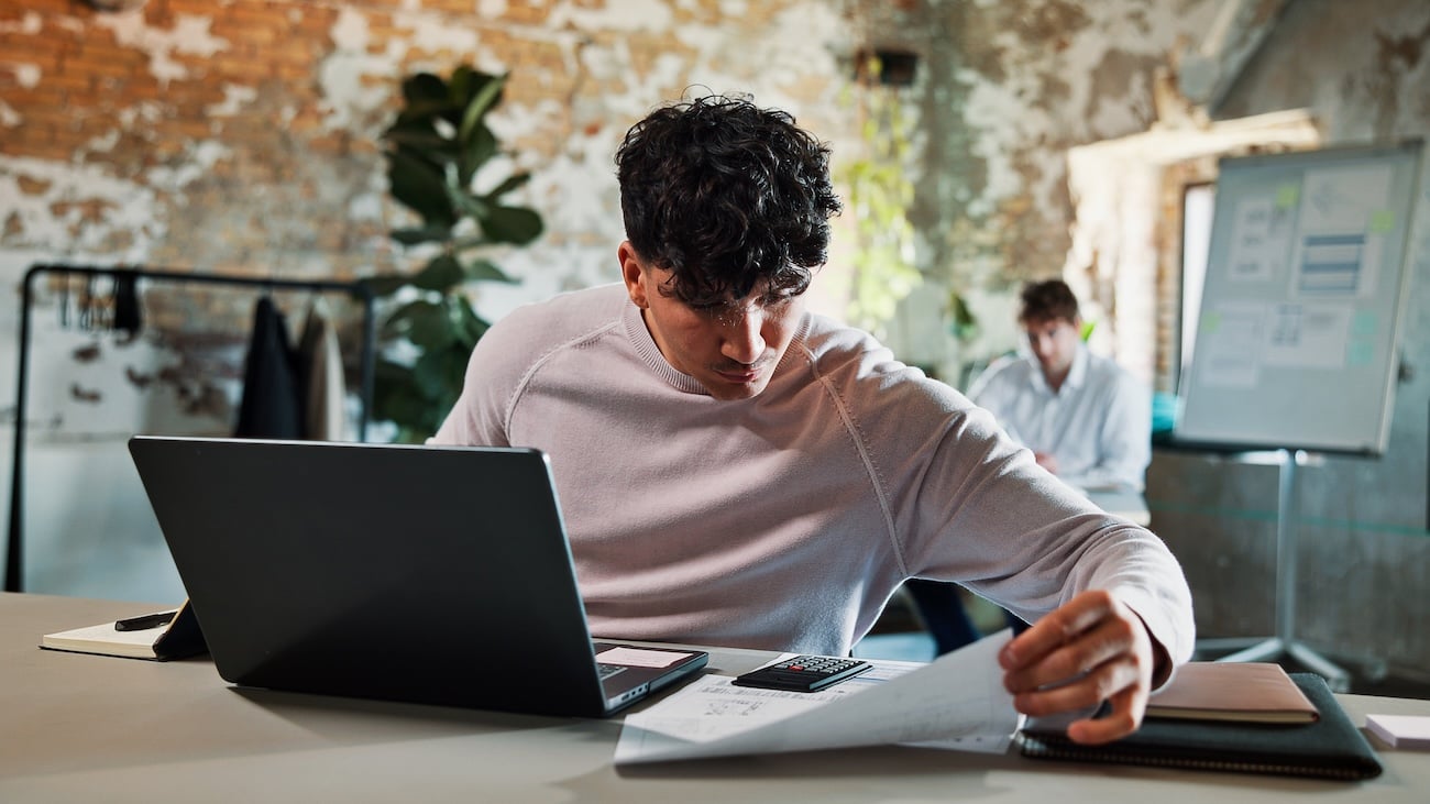 Ung mann sitter ved skrivebord med laptop og kalkulator og går gjennom dokumenter.