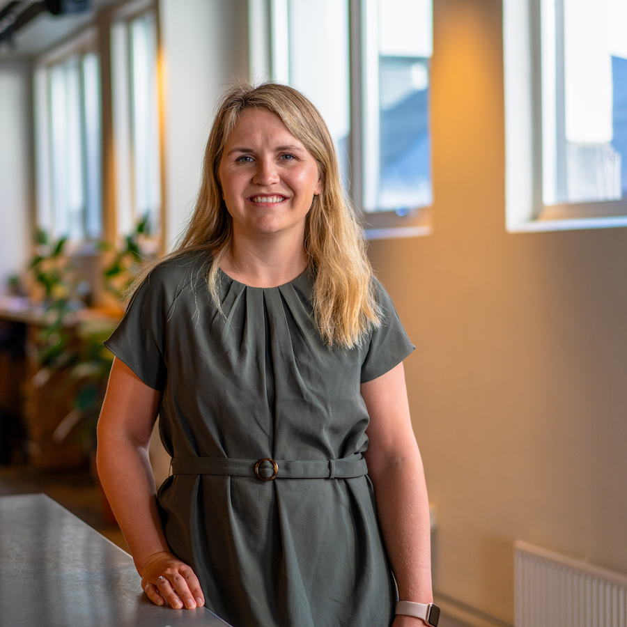Ung kvinne lener seg smilende mot en benk med vinduer uskarpt i bakgrunnen.