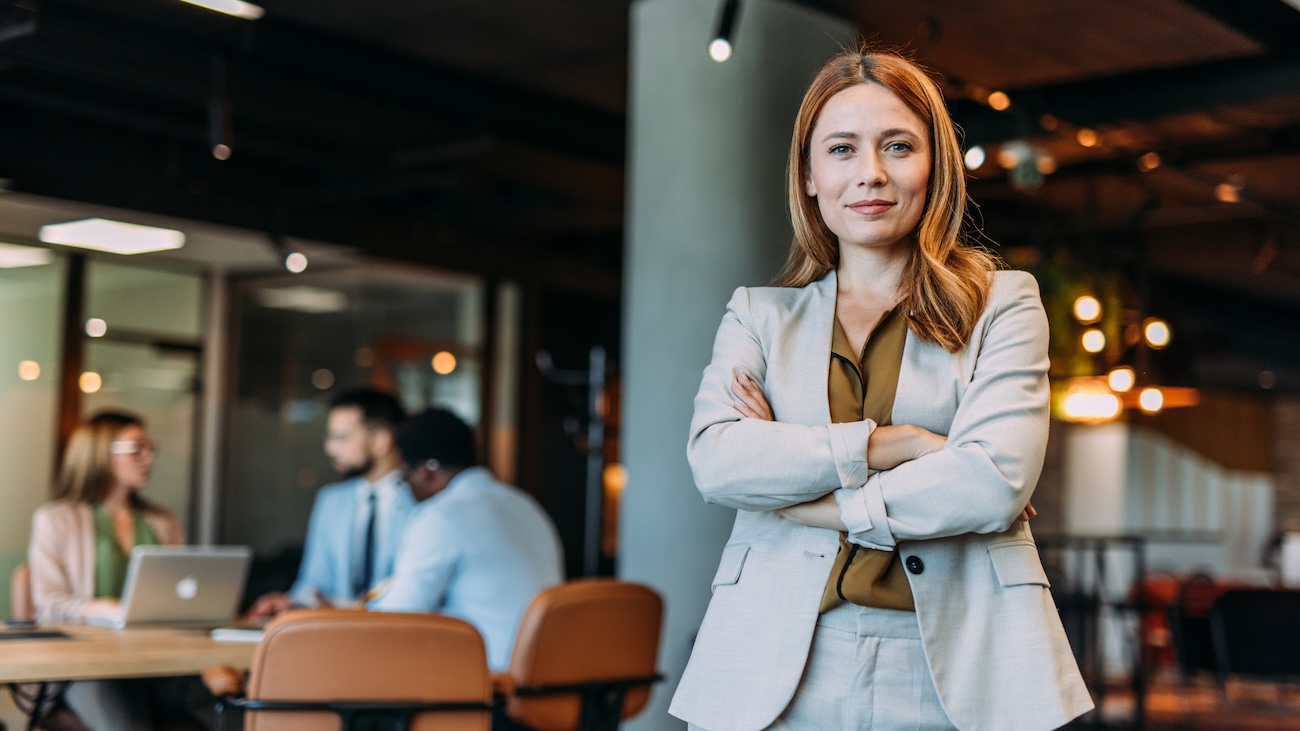 Formelt kledd kvinne som står i et kontorlandskap med armene i kryss mens hun ser mot kamera og smiler.