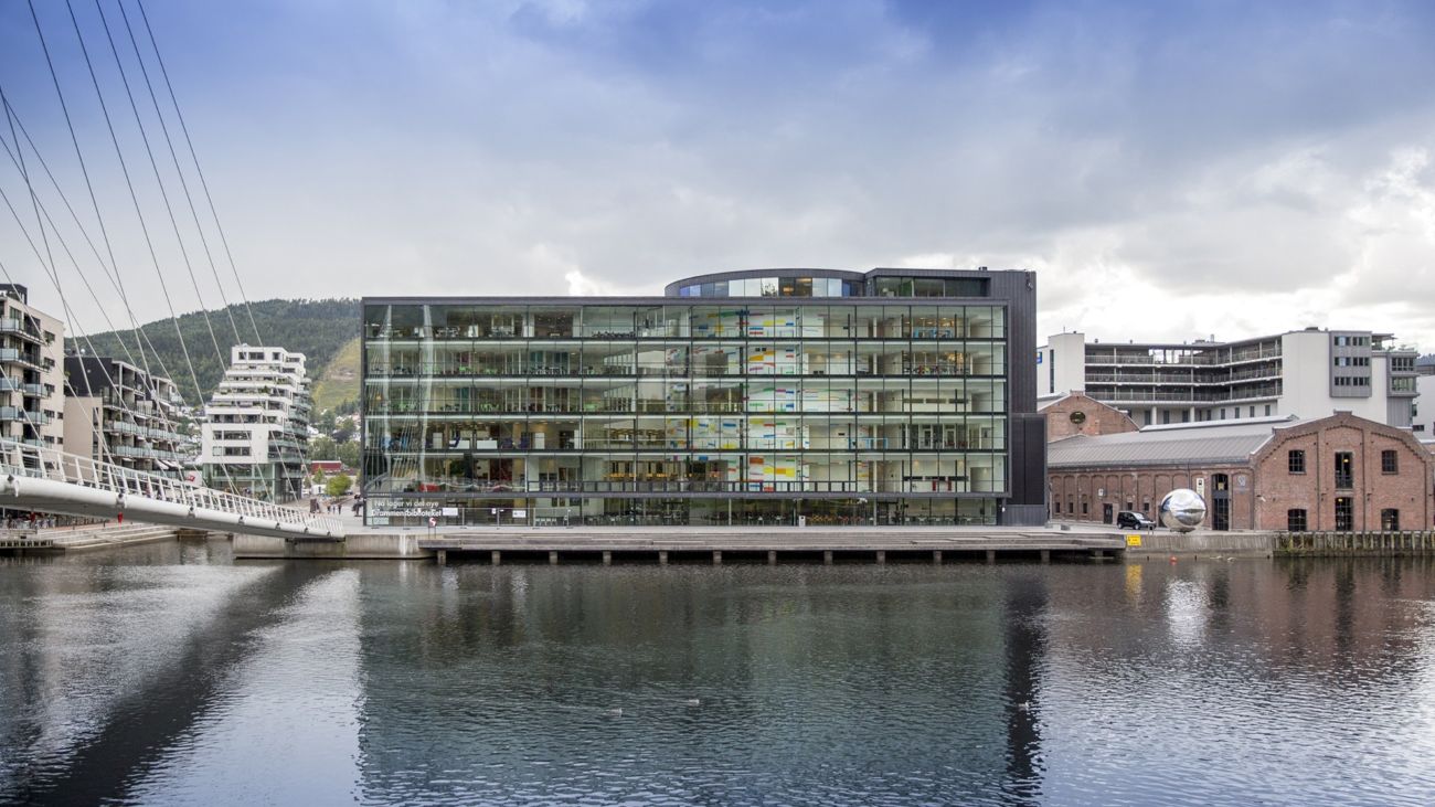  Moderne glasskontorbygning ved vannkanten med gangbro, som speiler seg i rolig vann. Overskyet himmel og synlig urban arkitektur.