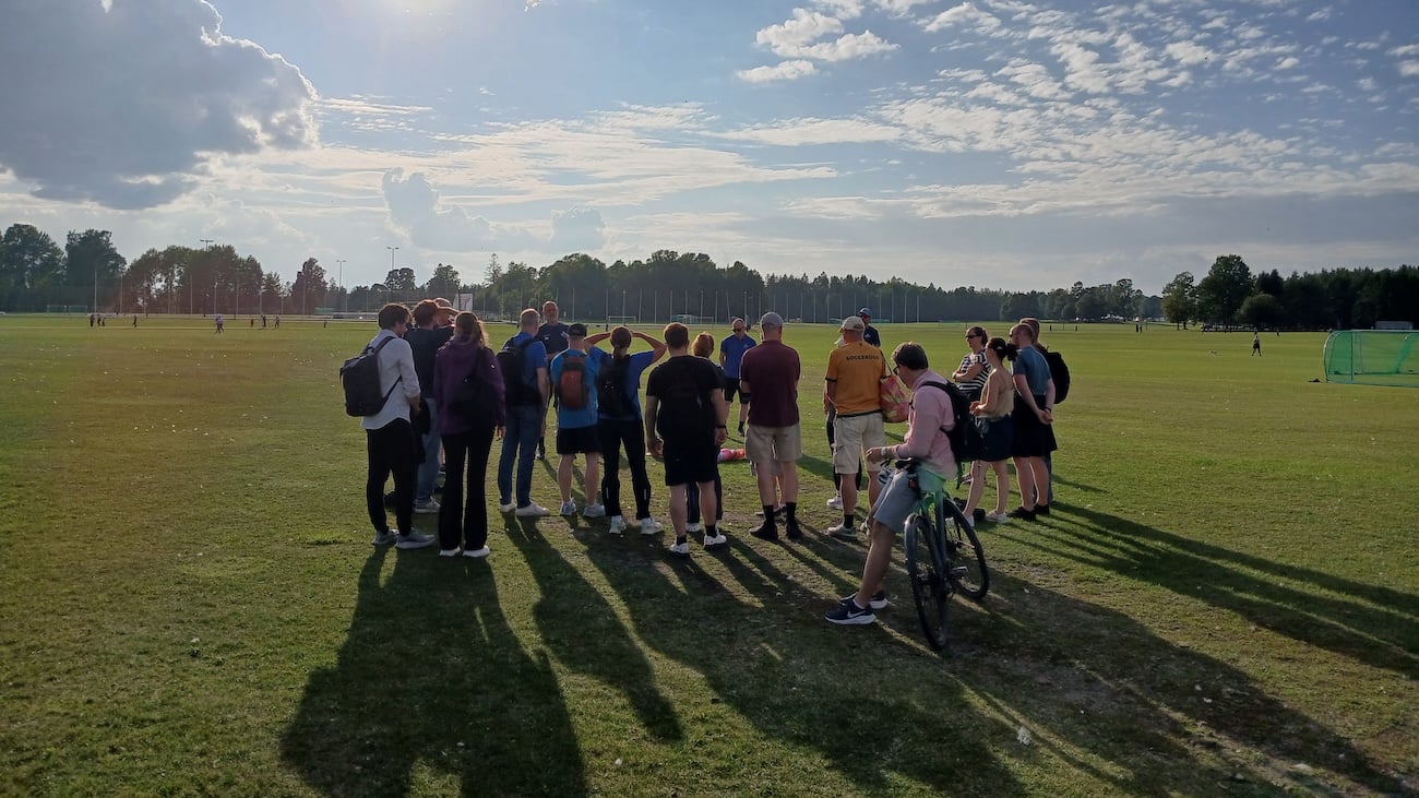 En gruppe mennesker står samlet på en gressbane i kveldssol, omgitt av trær og fotballbaner. En sykkel står foran gruppen.