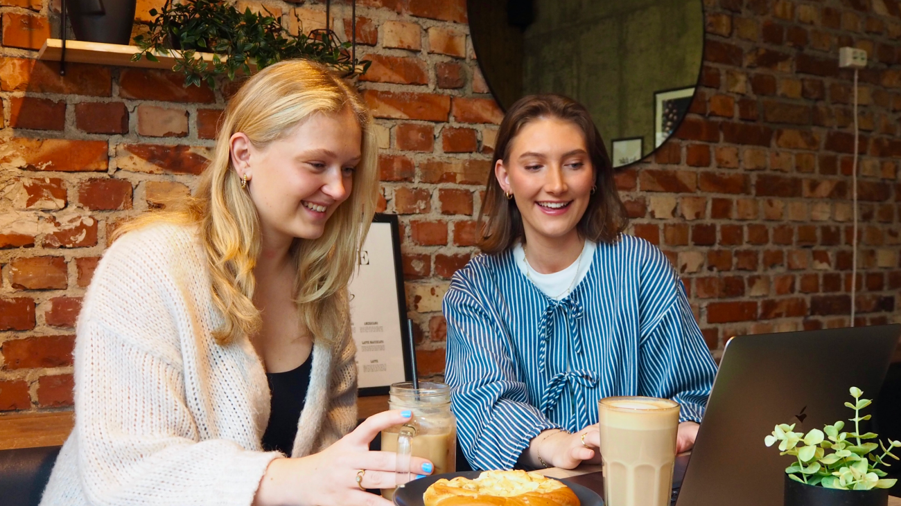 To unge kvinner som sitter sammen på en kafè og smiler mens de ser på en PC-skjerm.