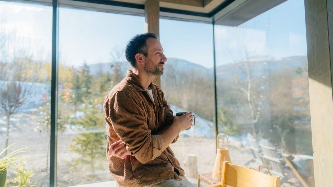 Mann som sitter i en hytte med store vinduer og nyter kaffen sin mens han ser ut mot vinterlige omgivelser.