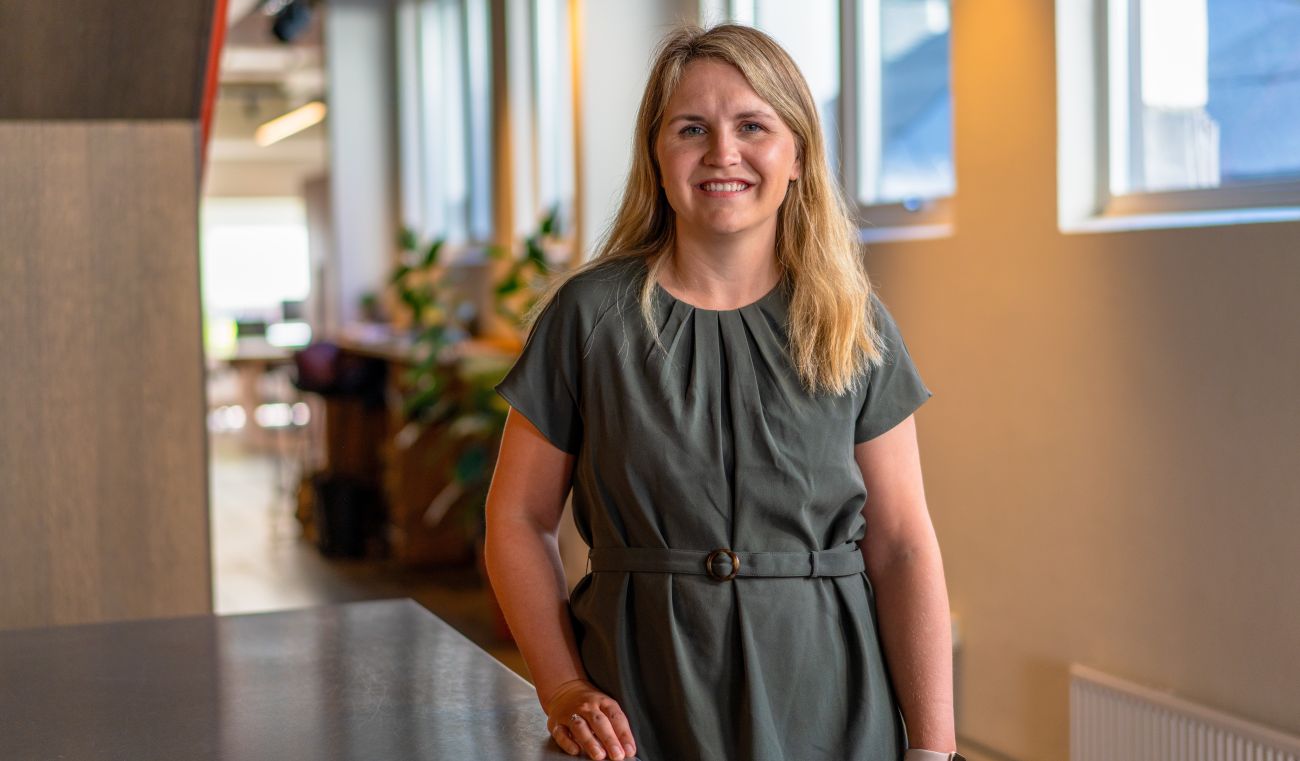 Kvinne som lener seg mot et bord i et kontorlandskap mens hun smiler bredt til kamera.