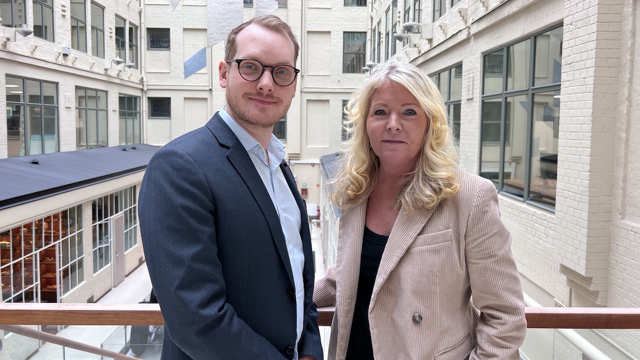 Advokatene i Econa Erlend Smith Eide og Anniken Astrup står ved et rekkverk i et lyst atrium med hvite murvegger og store vinduer.