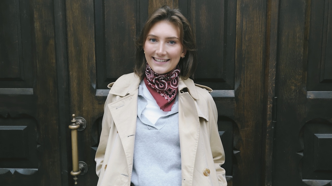 Nyutdannede Anja smiler foran en mørk tredør. Hun har på seg beige trenchcoat, grå genser og rødt paisleyskjerf.