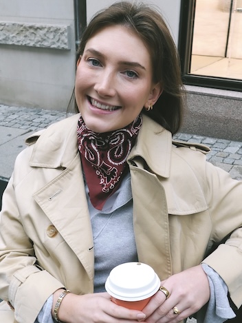 Nyutdannede Anja sitter ute i bygatene med en kaffekopp i hånden og smiler. Hun har på seg beige trenchcoat og rødt paisleyskjerf.