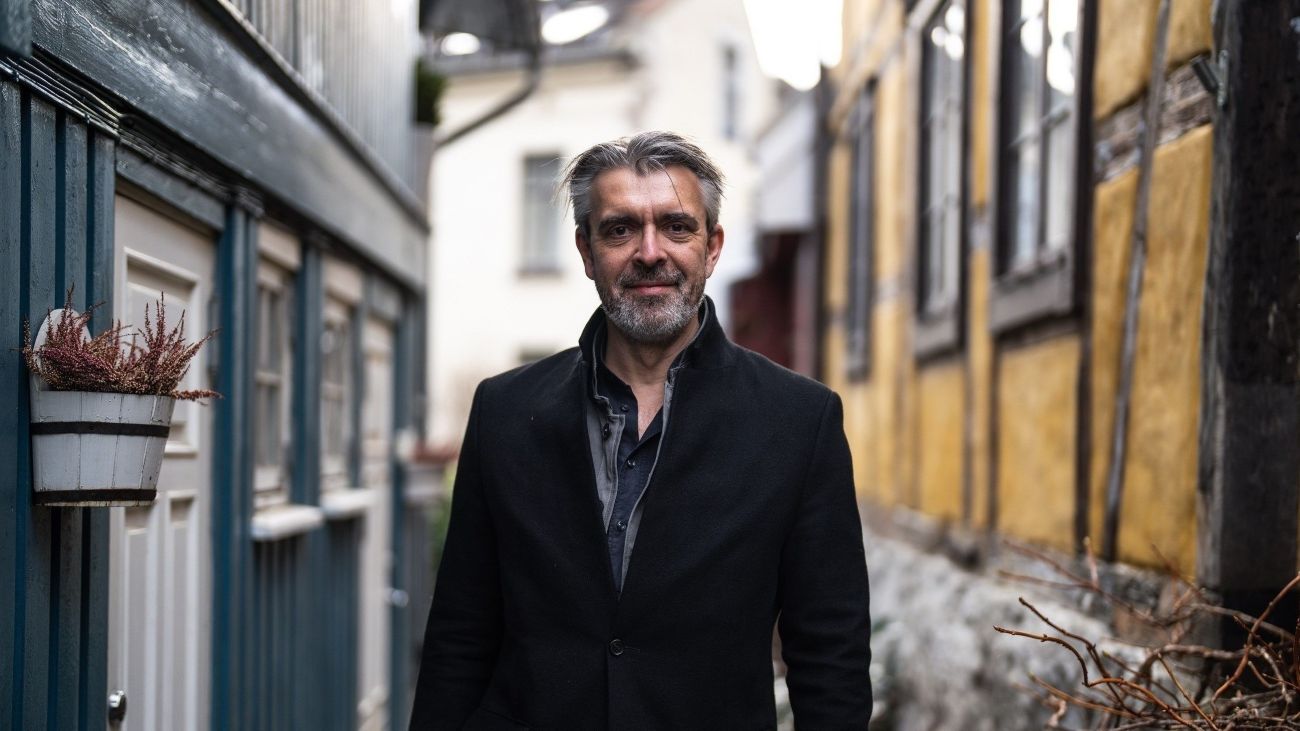 En formelt kledd mann som står mellom to hus i en smal gågate mens han smiler til kamera.