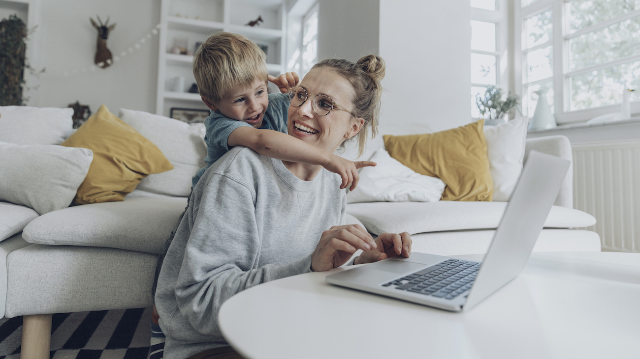 Mamma forran laptop på stuebord med ung gutt lenende over seg