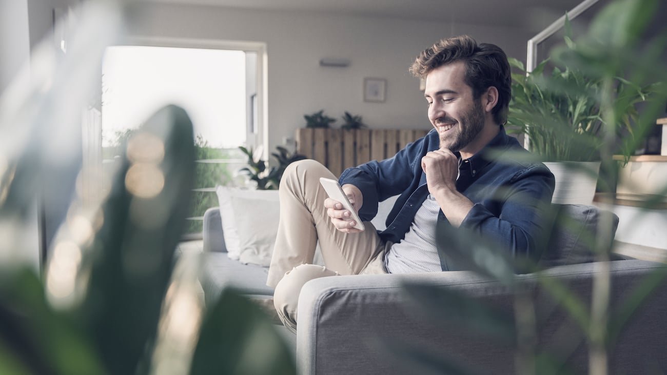 Smilende ung mann i sofa som ser på mobiltelefon