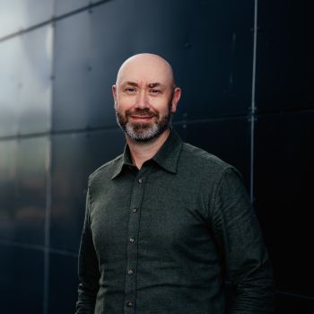 Portrett av mann som står utendørs foran en mørk vegg og smiler mildt til kamera.