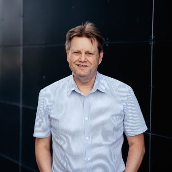 Portrett av en mann som står utendørs foran en mørk vegg, mens han smiler til kamera og holder hendene i lommene.