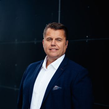 Portrett av formelt kledd mann som står utendørs foran en mørk vegg og smiler til kamera.