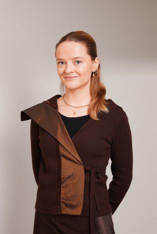 Portrettfoto av ung kvinne som står med hendene bak ryggen mens hun smiler forsiktig til kamera.