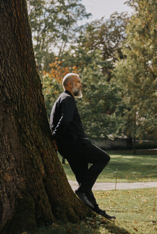 Mann kledd i sort som står og lener seg mot et stort tre i en park.