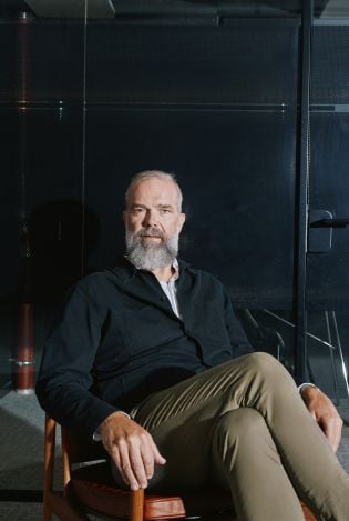 Mann som sitter lent bakover i en stol i et mørkt rom og ser rett inn i kamera.