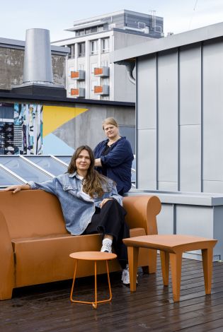 To kvinner i en brun sofa på en takterrasse som ser strengt i kamera.