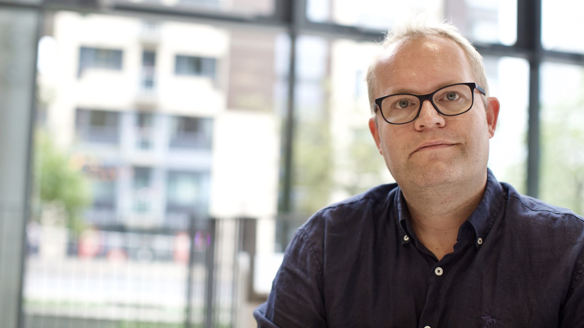 Mann som sitter i et lyst rom med store vinduer og ser rett inn i kamera.
