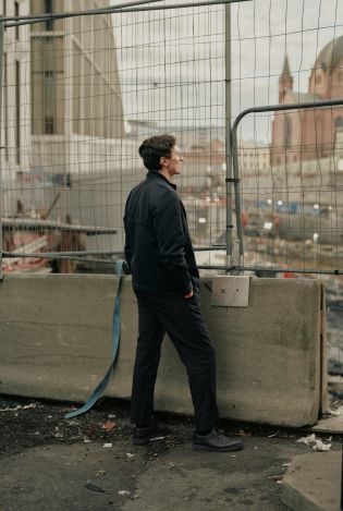 Mann som står foran gjerdene til en byggeplass og ser ut mot byen.