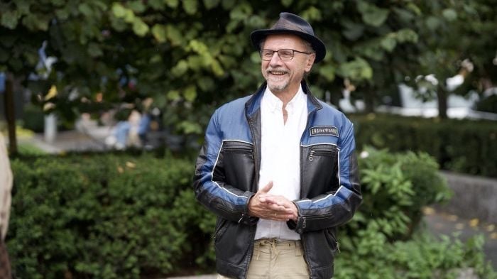 Stilig kledd mann som står ute i sommerlige omgivelser i byen, mens han smiler og gnir hendene mot hverandre.