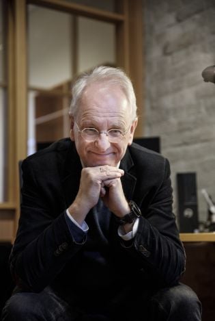 Mann som sitter og lener hodet på knyttnevene sine mens han lener seg mot kamera og smiler.