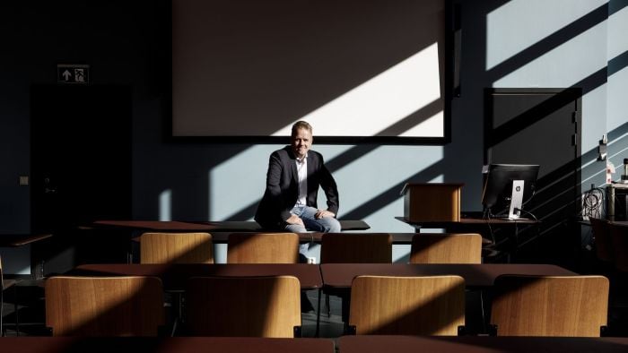 Mann som sitter på en pult i et mørkt undervisningsrom, mens han ser mot kamera og smiler.