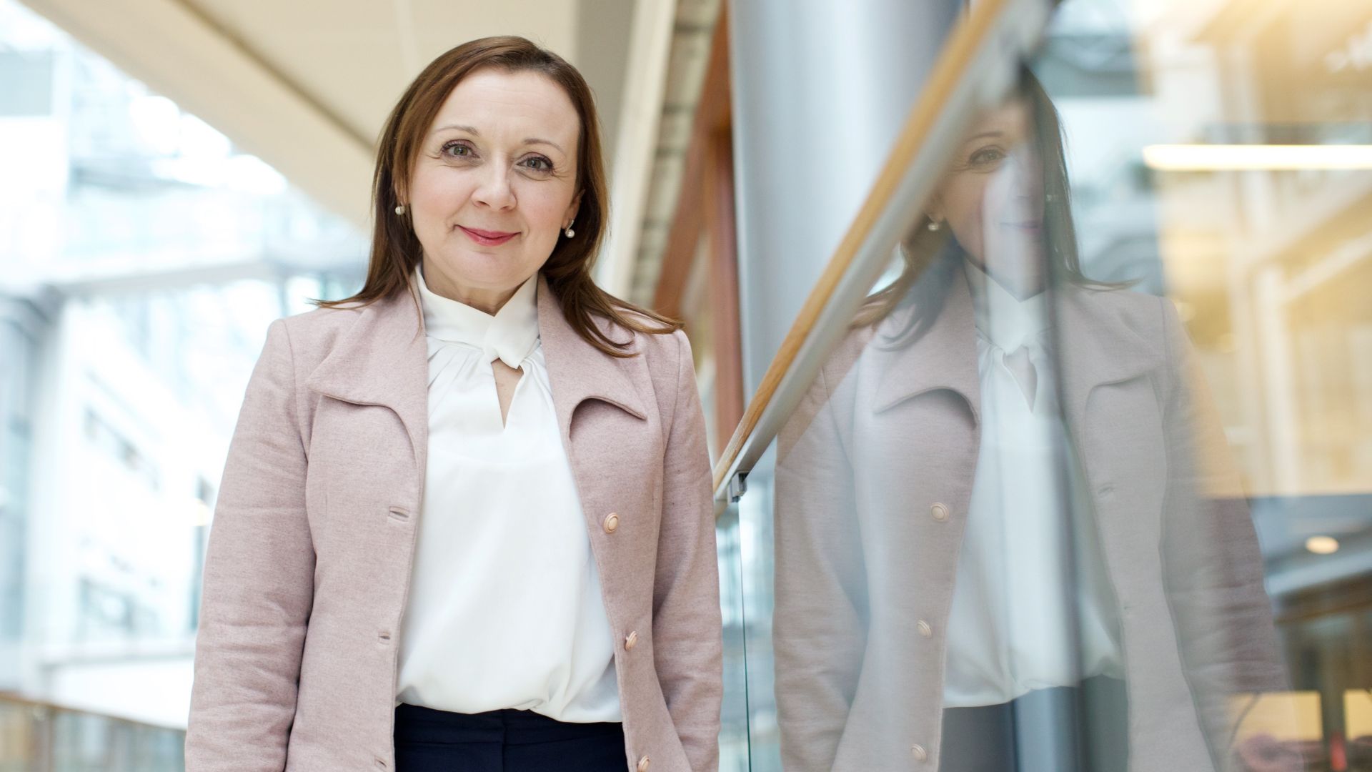 Kvinne som lener seg mot et glassrekkverk i et høyt og lyst bygg mens hun smiler til kamera.