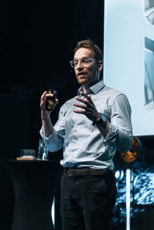 Mann som står på en scene foran en stor skjerm og holder foredrag.