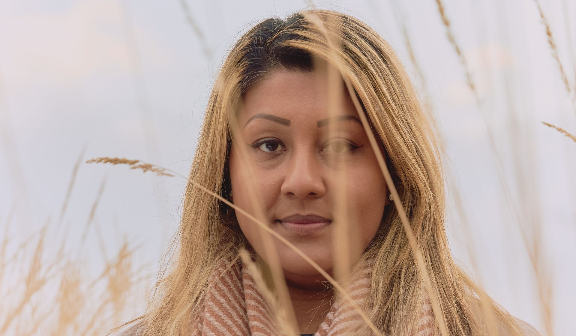Kvinne som står midt i en åker og ser frem mot kamera gjennom noen høye halmstrå.