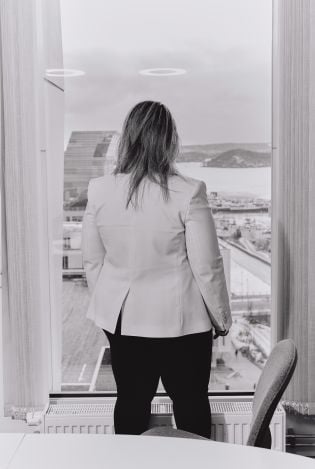 Svart-hvitt foto av kvinne som står med ryggen til kamera inne på et kontor mens hun ser utover Bjørvika gjennom vinduet.