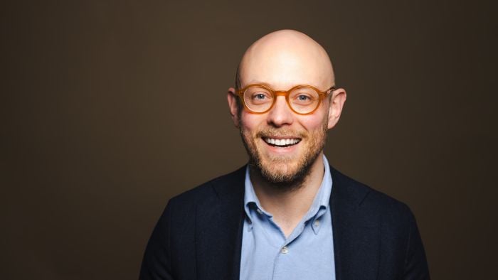 Formelt kledd mann som står i et mørkt rom og smiler bredt til kamera.