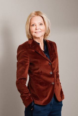 Kvinne i brun dressjakke i velour som står med hendene i lommene og smiler til kamera.