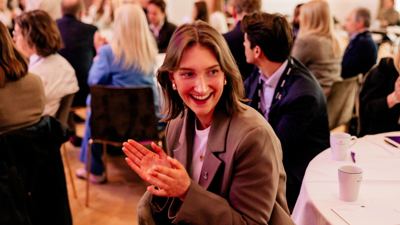 Ung kvinne som klapper og smiler mens hun sitter i en sal med mange mennesker rundt seg.